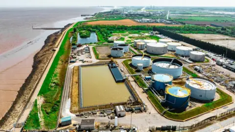 An aerial view of a biodiesel plant with a big area of water and circular buildings next to the beach and shoreline. 