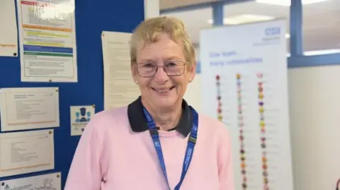 Sue Twose, a woman in her 70s, is wearing glasses and a pink jumper with a black collar. She is wearing a blue lanyard and is standing in a hospital corridor. 