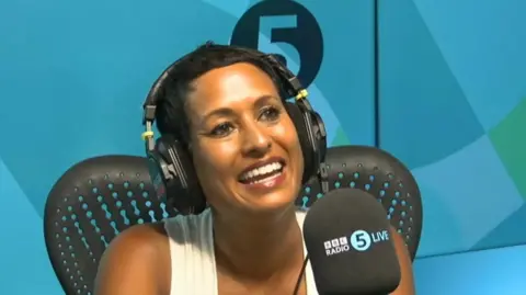 A woman with short, dark hair smiles as she talks into a microphone