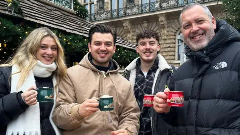 The Famileigh Darcy, Harrison, Travis and Paul, all holding mugs with drinks in them, looking at the camera and smiling. Darcy is wearing a white scarf and dark coat; Harrison a light-coloured coat; Travis a shirt and black coat; and Paul a black coat, done up.