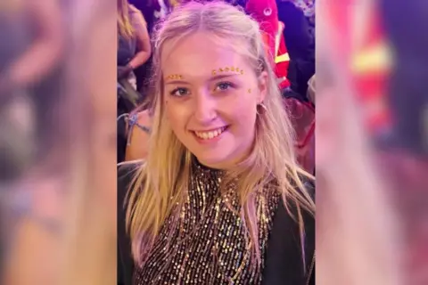 Meg Draper A woman has gold sequences above her eyebrows smiling at the camera. It is a head and shoulders shot. She wears a sparkly gold top.