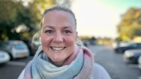 Jo Thewlis/BBC Charlotte Reed has long, blonde hair tied in a pony tail. She is smiling at the camera, wearing a cream jumper and a pink, blue and cream scarf wrapped around her neck. Behind her there are parked cars in the Butts Lane car park