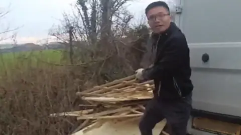A still of bodycam footage - Chunlin Zheng is next to the back of a white van holding some wood to put on a big pile of wood next to a hedge.