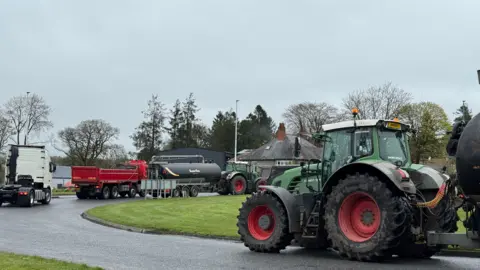 A number of tractors and trucks driving around a roundabout on a grey overcast day.