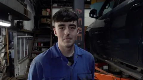 A man with brown hair which is long on the top and short at the sides looking into the camera and wearing a blue overalls shirt. He is standing in a mechanic's garage with various objects behind him and a black car to the right of him.