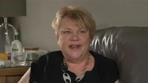 A woman with short blonde hair wearing a black T-shirt and a necklace smiling in the direction of the camera