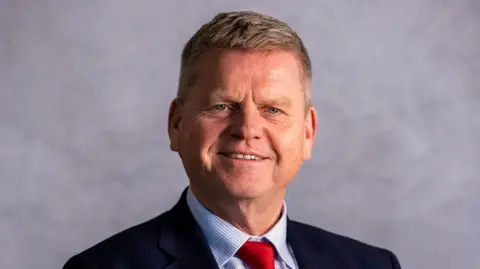 A picture of Prof Robert Van de Noort, who is stood in front of a grey background. He is wearing a dark blue jacket, blue and white checked shirt and a red tie.