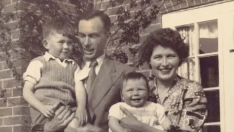 Barbara Forey A black and white photo from the early 1950s showing L to R, Robert Bert Bowman, Barbara and Eileen Bowman outside a house. Robert is about four and is held up by his father, who is wearing a suit and tie. Barbara is about one and is being carried by her mother, in a patterned dress. 