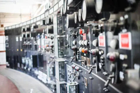 A long wall of control panels, with buttons, dials, lights and switches