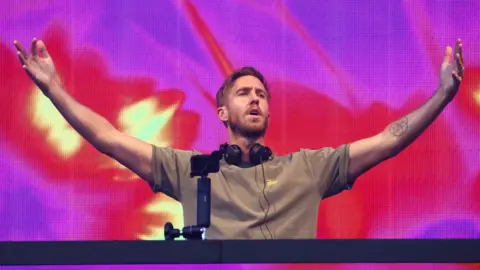 Calvin Harris wears a plain grey T-shirt and stands at the DJ booth on stage. He has headphones around his neck and holds his hands up in the air. Behind him you can see a screen with purple, red and yellow shapes 