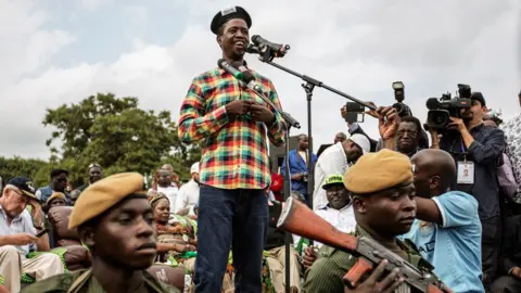 AFP via Getty Images Edgar Lungu speaks at a campaign rally. Dressed in a chequered shirt and beret he is surrounded by photographers and soldiers stand in front of him.