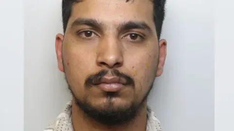 A man with a dark moustache and hair in police custody.