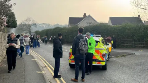 Crowds of people mill around on a busy street outside Weston hospital. A police car monitors the scene. Smoke can be seen in the background.
