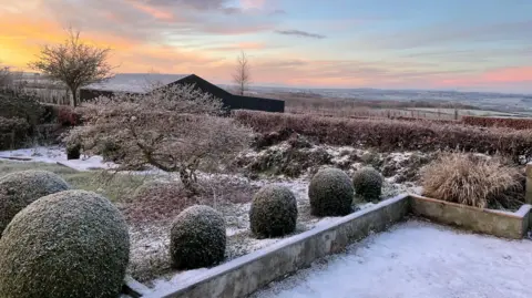 BBC Weather Watchers/BeechyJ The image shows snow and frost in a garden. Snow has settled on what appears to be a patio area and on an area of grass and small bushes. 