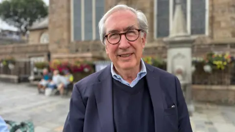 A elderly man with grey hair and round glasses smiling at the camera. He is wearing a dark blue suit jacket, with a dark blue v neck jumper underneath. You can see the collar of a light blue shirt with thing white stripes.