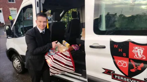 Family A teenager in a black school uniform holds a red and white striped gift bag with presents in it, which he takes out of a white school van with a black and orange logo on the door