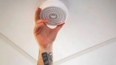 A man installing a vape detector in a school building, with his arm reaching up to the ceiling to connect the device. 