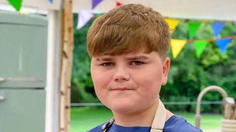 A headshot of a boy with brown hair, blue t-shirt and sand apron. He is standing in a marquee with colourful bunting and a green fridge.