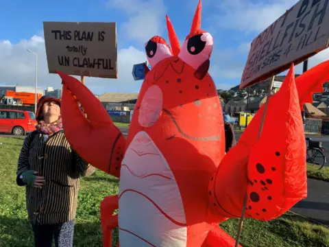 Someone in a lobster costume holding a placard saying 'don't be shellfish, clean seas for all', and a woman to the side holding a placard saying 'this plan is totally unclawful'