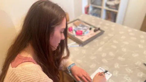 Lara is sitting at a table writing a note. The tablecloth has a faint grey and white dandelion pattern. 