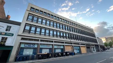 The front of the Thomson House building, which is partly boarded up at ground level. Signage for the Metro, Chronicle and Sunday Sun newspapers is in some of the windows, along with graffiti. A number of wheelie bins are on the pavement.