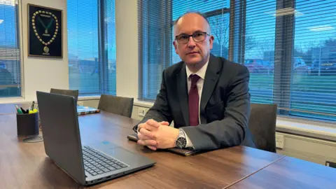 A man wearing a grey suit, white shirt and burgundy tie sits at a brown desk with his hands clasped together. He is looking towards the camera. In front of him is an open laptop. He is sitting in an office surrounded by windows