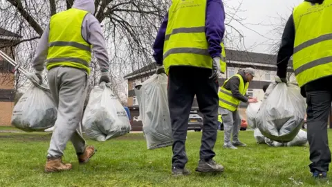 Dan Bater/BBC A group of people wearing casual clothes and high-vis yellow jackets. They are on a patch of grass and are holding bags of rubbish in their hands.