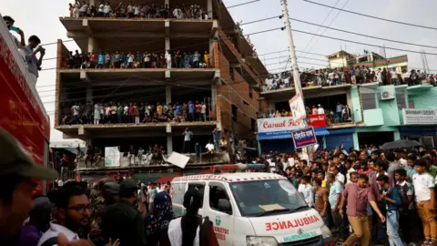 Reuters Crowds of people stand on the streets and on unfinished buildings looking down at an ambulance which makes its way through the crowd.