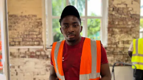 Brandon is wearing an orange high-vis jacket, red t-shirt and looking straight at the camera.