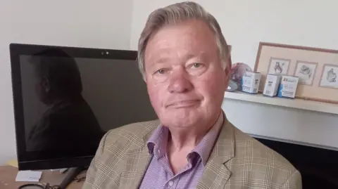 Supplied A photo of Hamish Brown. He is wearing a blazer over a shirt. He is sitting in front of a computer screen but is looking directly into the camera. 