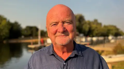 Martin Stewart smiles for the camera. He has a grey stubble beard and is wearing a dark blue open-collar shirt