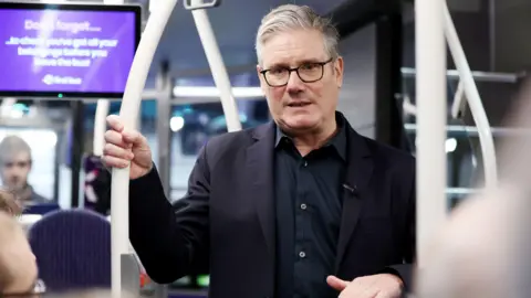 BBC Sir Keir Starmer stands with his hand holding a rail in a bus. He is wearing a dark shirt and jacket and has black-rimmed glasses on. He has grey hair and is speaking to someone sitting down. 