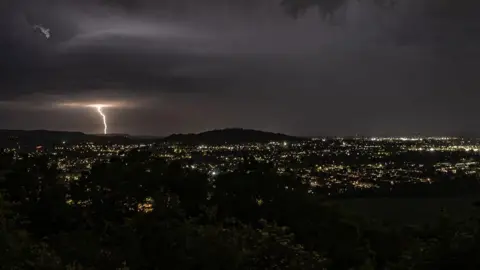 Damian Conner-Smith A gloomy night sky is lit by a bolt of lightning in the distance. City street lights are dotted all over the land in the foreground. 