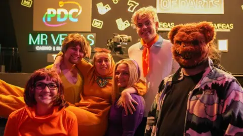 City of Wolverhampton Council Six people are in the photo, including someone on the right with a bear face mask on. The woman who is the third person from the left has her arms around the two people either side of her.