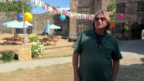 A man wearing a green shirt stands in a castle courtyard decorated with colourful bunting and hanging lanterns.