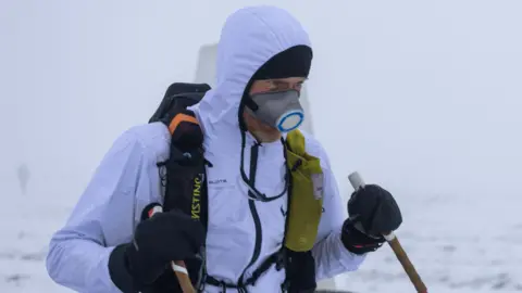A man in a white waterproof jacket with the hood up, standing in a snowy landscape. He is also wearing a backpack and a mask over his face, and is carrying walking poles.