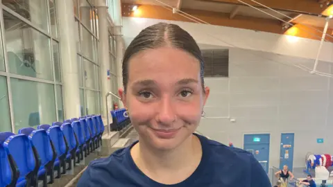 Olivia Newman-Baronius, who is wearing a blue top, smiles in a picture taken at the side of a swimming pool 