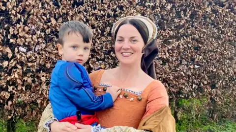 Supplied Hollie dressed in a medieval costume as she holds her son Max in her arms. Max is also wearing a costume, with a blue long-sleeved top and red belt.
