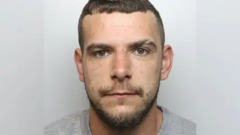 A police mugshot of Gregory Twigg, he is stood against a light-coloured backdrop.