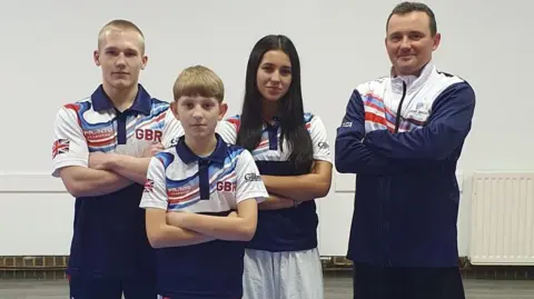 Three teenagers wearing Team GB kit are posed with their arms crossed. They are standing next to a man who also has his arms crossed. 