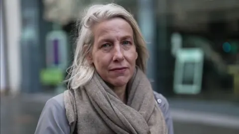 A head and shoulders shot of Helen Dickinson looking straight at the camera. She is standing outside on a dull winter day, wearing a grey coat and a large grey scarf.  