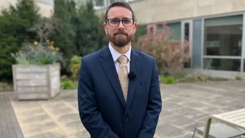 Robert Smith is a bespectacled man with dark hair and neat beard. He is wearing a dark blue suit, white shirt and gold toe and is standing outside a modern, blonde sandstone building. 