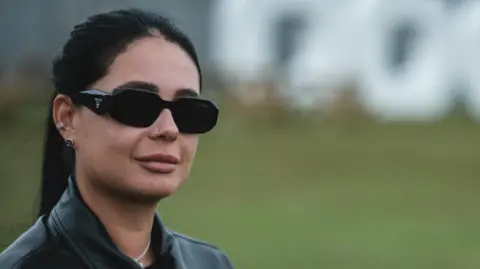 Hannah Laing, in dark sunglasses, her dark hair in a ponytail and wearing a black leather jacket, stands with a large "Doof" sign behind her in Camperdown Park