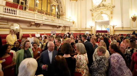 Reuters A huge crowd of people in an ornately-decorated room with what looks like a pipe organ on a balcony. King Charles is in the midst of the crowd with a drink in his hand as he talks to people.