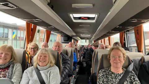 Shariqua Ahmed/BBC A group of people siting inside a bus with their seat belts fastened. They are all smiling at the camera. 