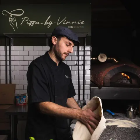 Pizza by Vinnie A man wearing a black chef's uniform and a cap holds pizza dough in his hand. A pizza oven is on the surface next to him and a green sign on the wall says 'Pizza by Vinnie'.