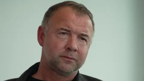 A man, who is slightly frowning, looks to the right of the camera. He has thin hair with some grey in it. He wears a denim jacket with a black T-shirt underneath.