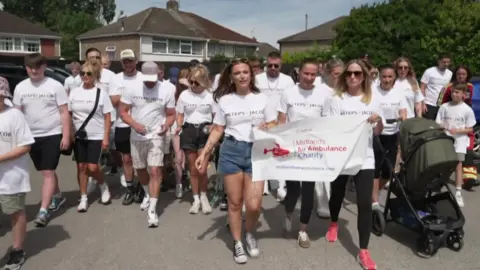 A group of people in white t-shirts take part in a walk, starting in a car park with houses visible behind them.