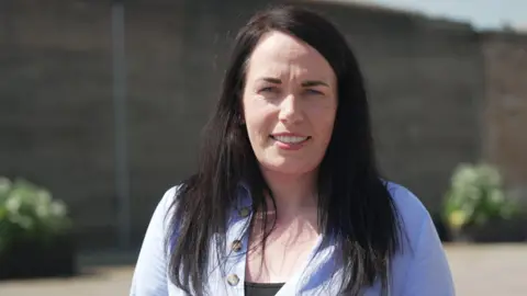 A close-up outdoor portrait of a woman facing the camera. She has long dark hair and is wearing a light blue buttoned top over a darker underlayer.
