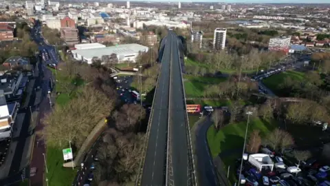 BBC An aerial photograph of the Gateshead flyover. The bridge on the A167 runs towards Gateshead town centre and the Tyne Bridge. A car dealership can be seen to the right of the bridge while commercial buildings stand to the left. Newcastle can be seen in the distance.